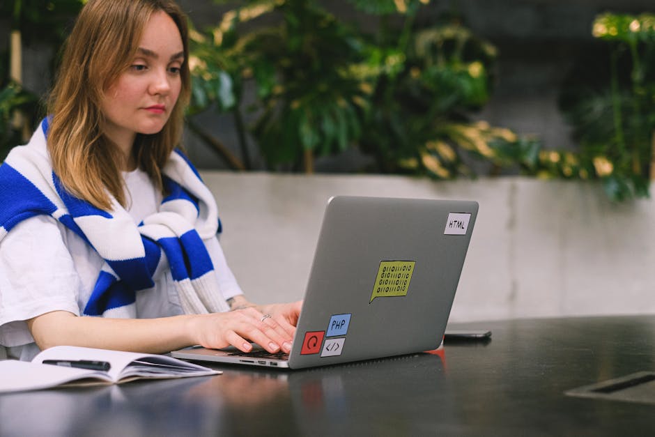 A young woman with shoulder-length light brown hair sits at a black outdoor table in a garden setting, working on a silver laptop that displays various colorful stickers, including one labeled 'HTML,'